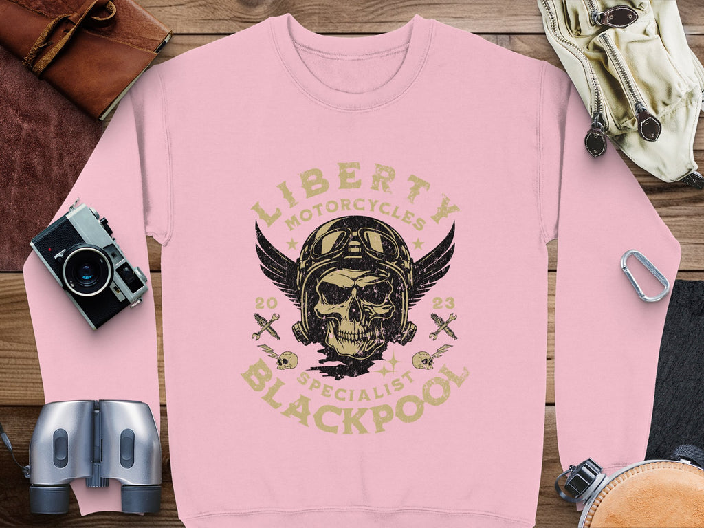 A pink sweatshirt by Garment Graphics showcases a skull in a helmet with wings and crossbones, surrounded by Liberty Motorcycles Specialist Blackpool 2023. Perfect for the biker look, its placed on wood next to binoculars, a camera, gloves, and a leather pouch.