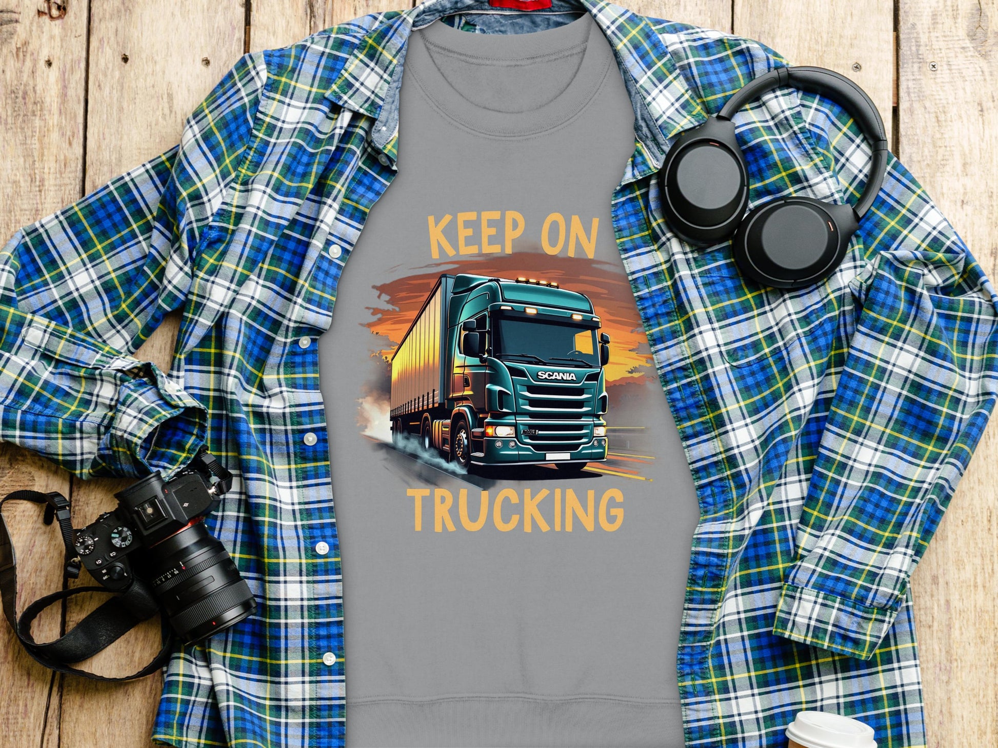 Garment Graphics gray t-shirt, adorned with a vibrant truck graphic and Keep On Trucking text, lies on wood. Its accompanied by a blue plaid shirt, headphones, a camera, and a white coffee cup—ideal for truck enthusiasts celebrating every mile.