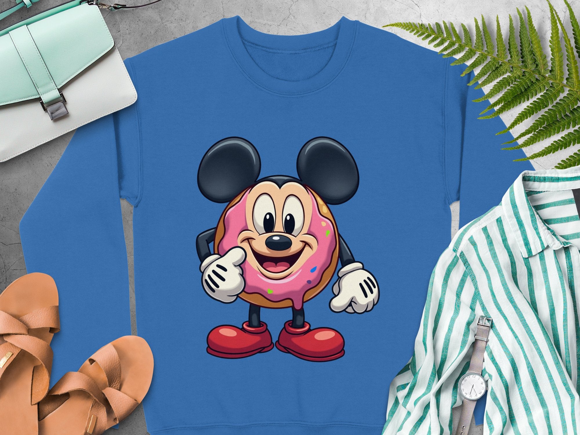 Garment Graphics presents a unisex blue sweatshirt with a charming donut-bodied mouse in pink frosting, hands on hips. Nearby are a green striped shirt, brown sandals, a wristwatch, and a teal purse among lush fern leaves.