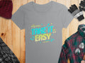 The gray T-shirt from Garment Graphics boasts vivid graphics and the phrase Take It Easy with bright colors, arrows, and musical notes. It lies on a wooden surface accompanied by a plaid shirt, knit gloves, and a wool hat for a stylish ensemble.