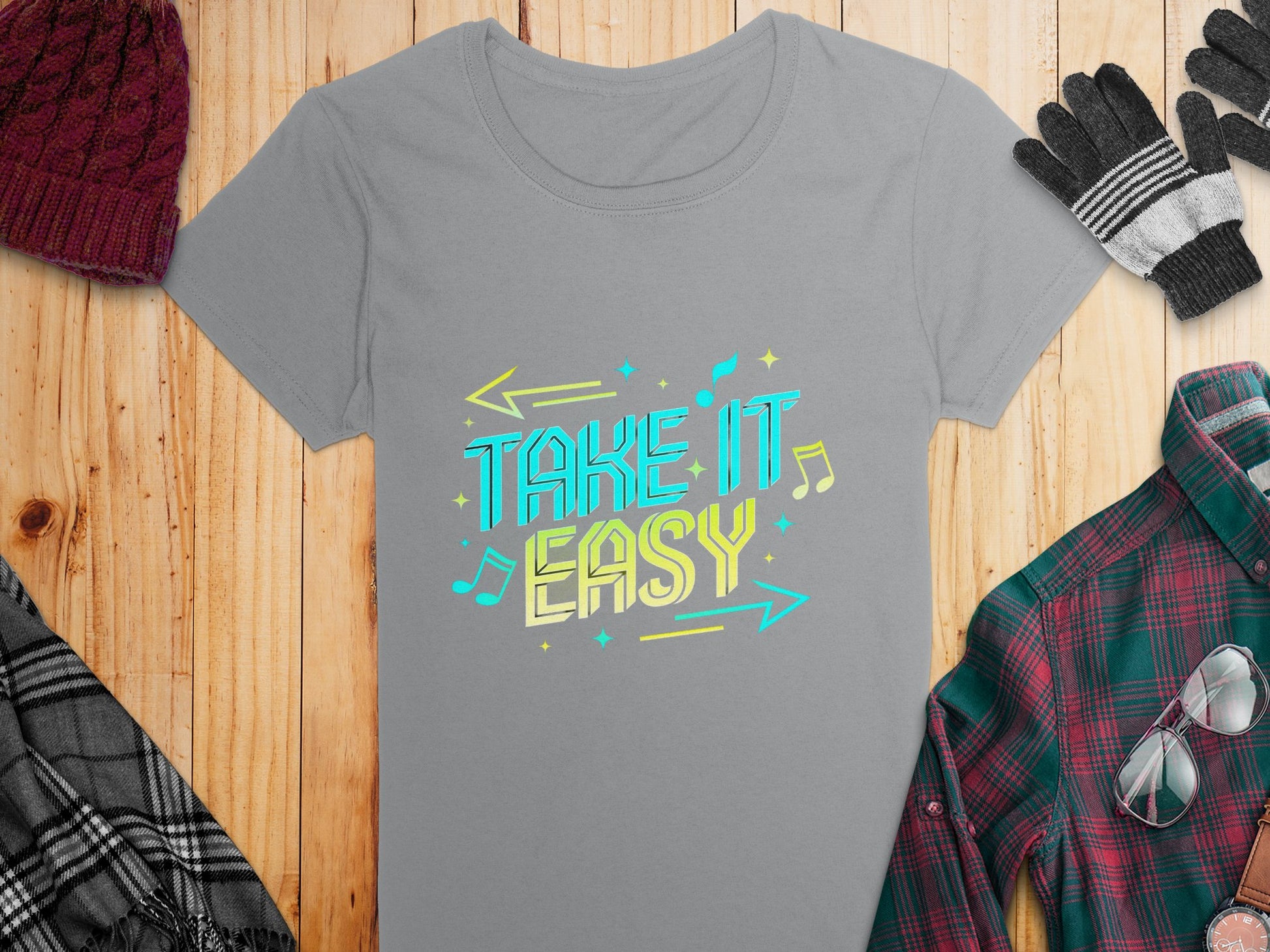 The gray T-shirt from Garment Graphics boasts vivid graphics and the phrase Take It Easy with bright colors, arrows, and musical notes. It lies on a wooden surface accompanied by a plaid shirt, knit gloves, and a wool hat for a stylish ensemble.