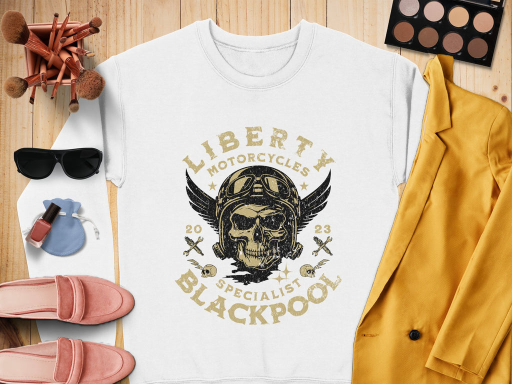 Flat lay of Garment Graphics white sweatshirt with a skull and Liberty Motorcycles Blackpool print, ideal for a biker look. Features include yellow jacket, pink shoes, sunglasses, makeup palette, brushes, nail polish, and a small blue pouch on wooden surface.