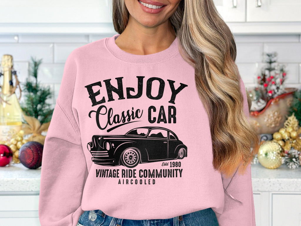 In a festively decorated kitchen, a woman wearing a pink sweatshirt from Garment Graphics, featuring Enjoy Classic Car, Vintage Ride Community and a charming car graphic, stands proudly. A basket of ornaments enhances the nostalgic ambiance of her vintage-inspired attire.