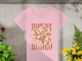 A pink womens Rodeo T-shirt from Garment Graphics features a cowgirl on horseback with Howdy Rodeo and Not Beginner in a badge shape. It rests on concrete, framed by green and yellow plants.