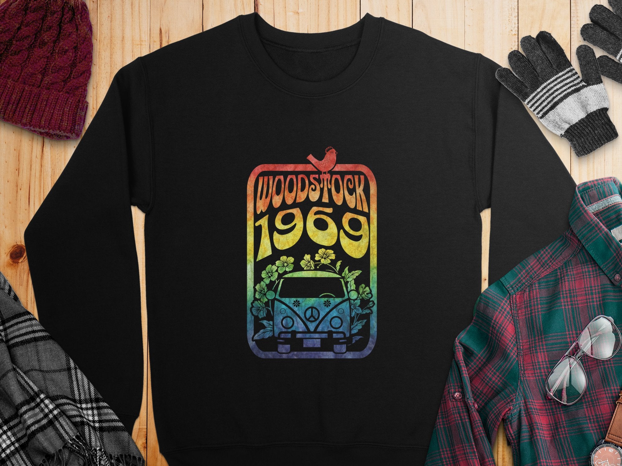 A black Garment Graphics sweatshirt features a vintage van graphic with flowers, a bird, and Woodstock 1969 in rainbow text. Evoking retro style, its displayed on wood beside a red knit hat, gloves, and a plaid shirt.