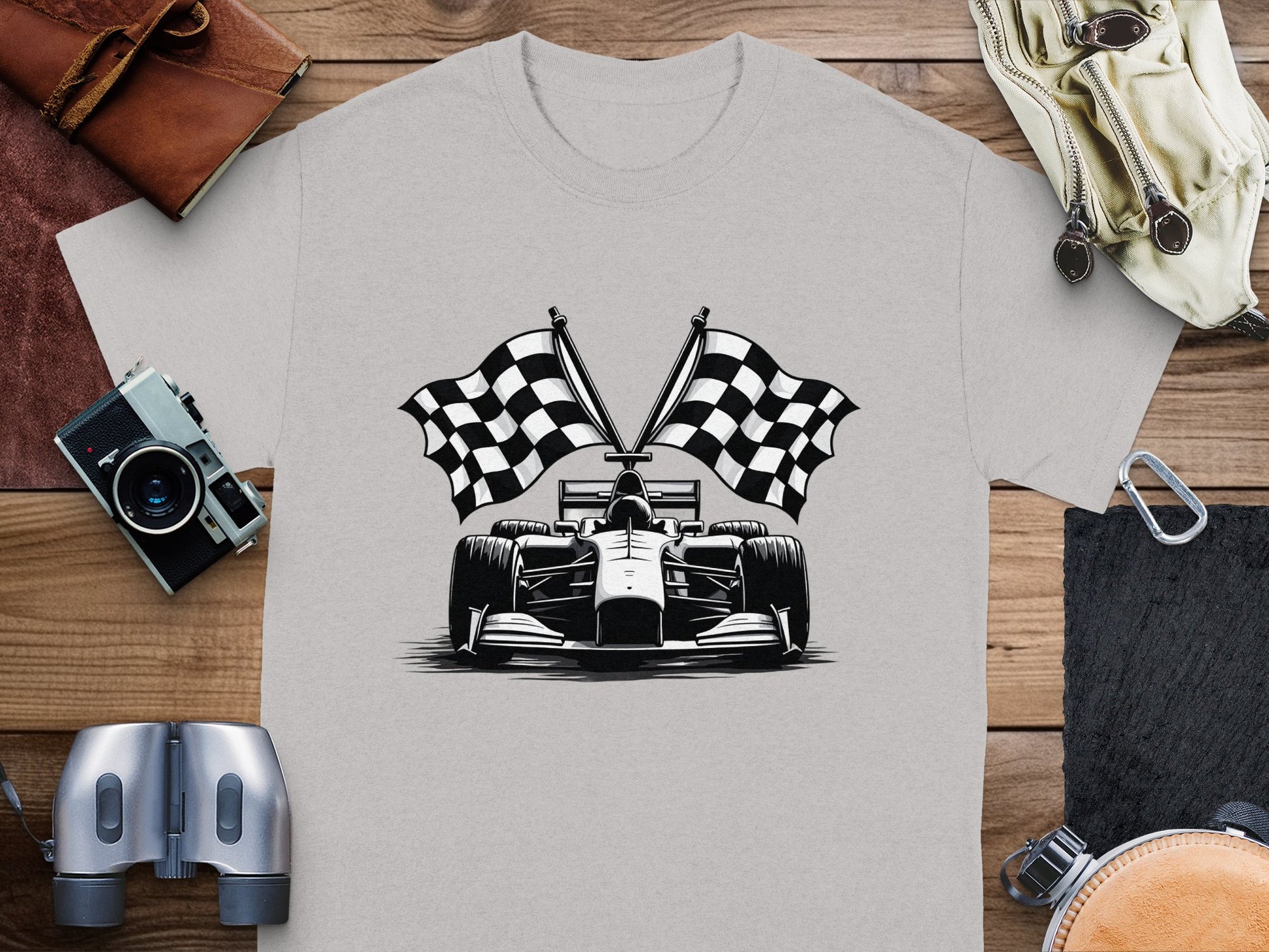 Garment Graphics gray T-shirt features a black and white Formula 1 car illustration with crossed checkered flags. Its displayed on wood, surrounded by a camera, brown leather pouch, binoculars, and beige bag—ideal for motorsport fans.