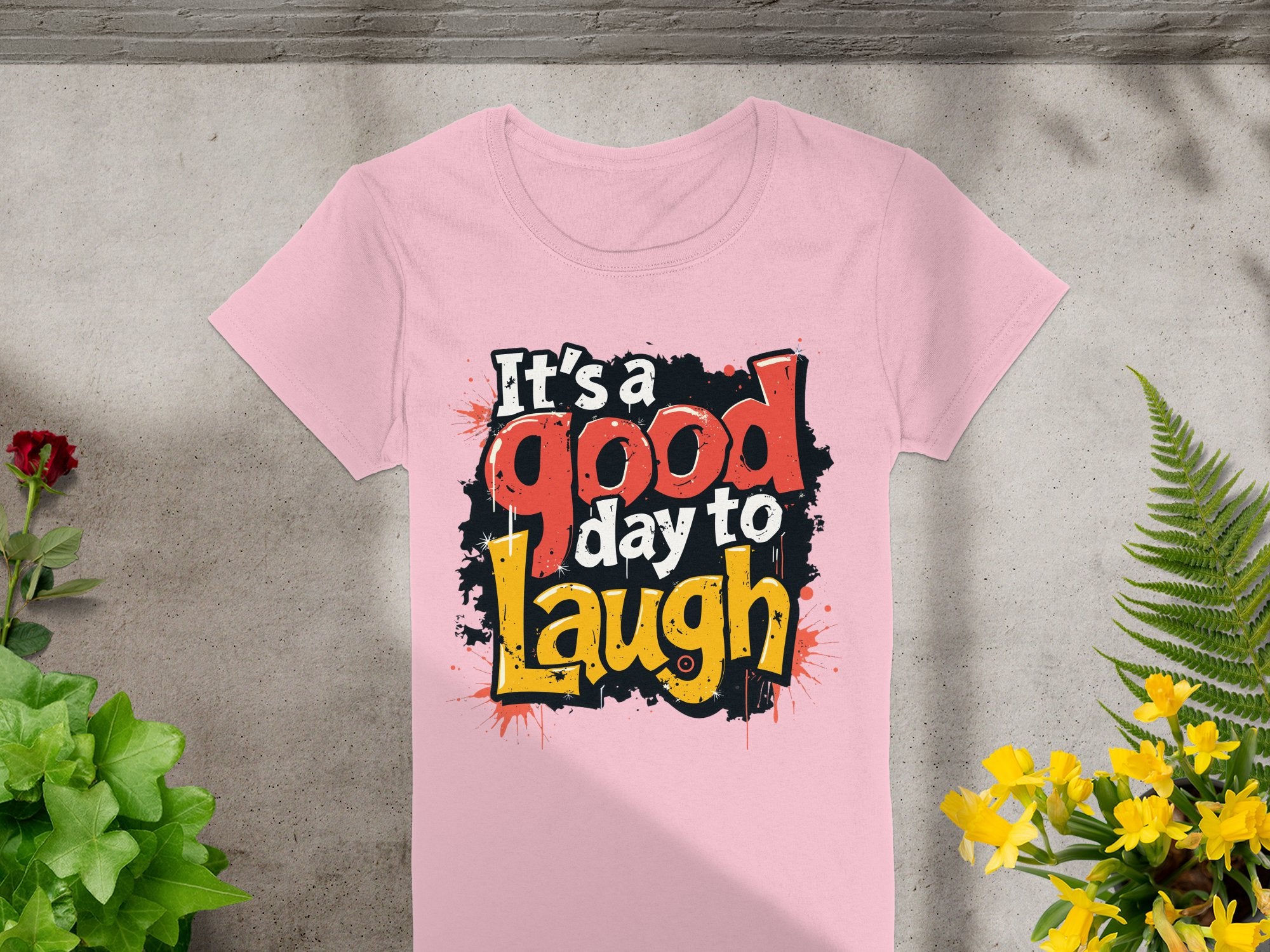Garment Graphics semi-fitted pink t-shirt features bold, stylized Its a good day to laugh text on a textured gray background with vibrant red and yellow flowers and lush green foliage for a positive touch.