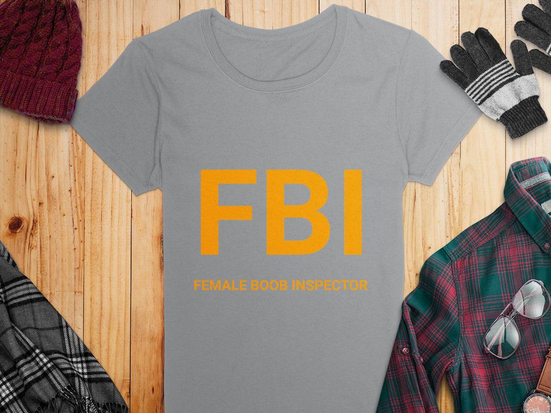 A white Garment Graphics T-shirt features yellow FBI Female Boob Inspector text, lying on wood. Its surrounded by holiday items: gifts, a deer-patterned sweater, pine cones, red ornaments, and a candy cane. Ideal as a gag gift and available in womens sizes.
