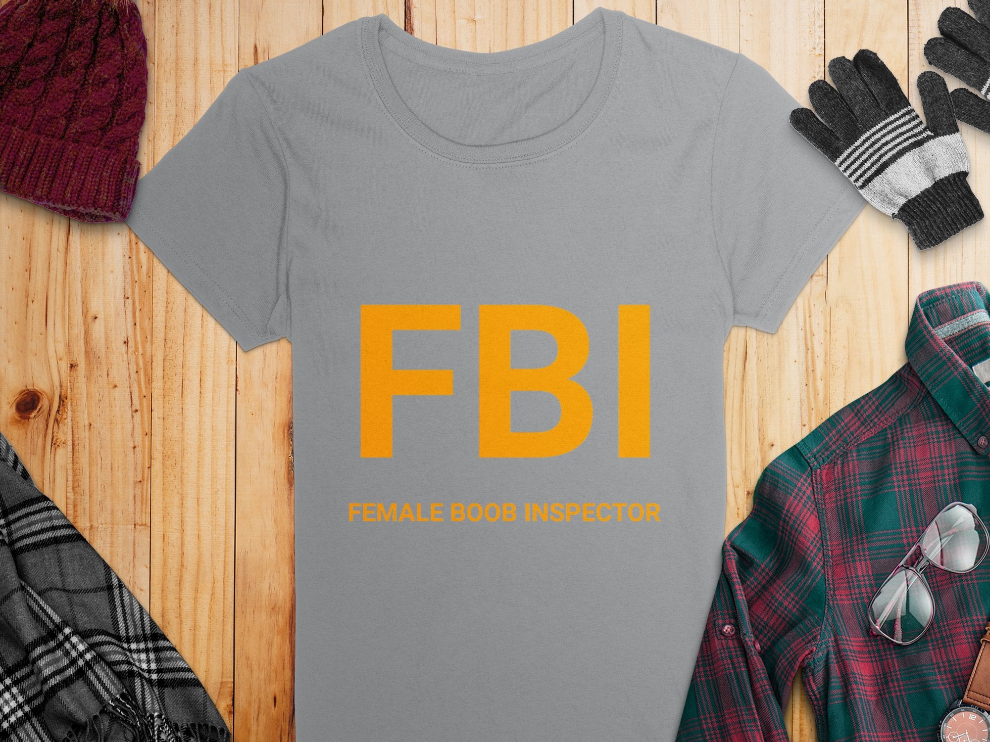 This Garment Graphics gag gift, a womens gray FBI Female Boob Inspector T-shirt, features bold orange letters and smaller text. Its pictured on wood with a plaid shirt, gloves, and knitted hat.