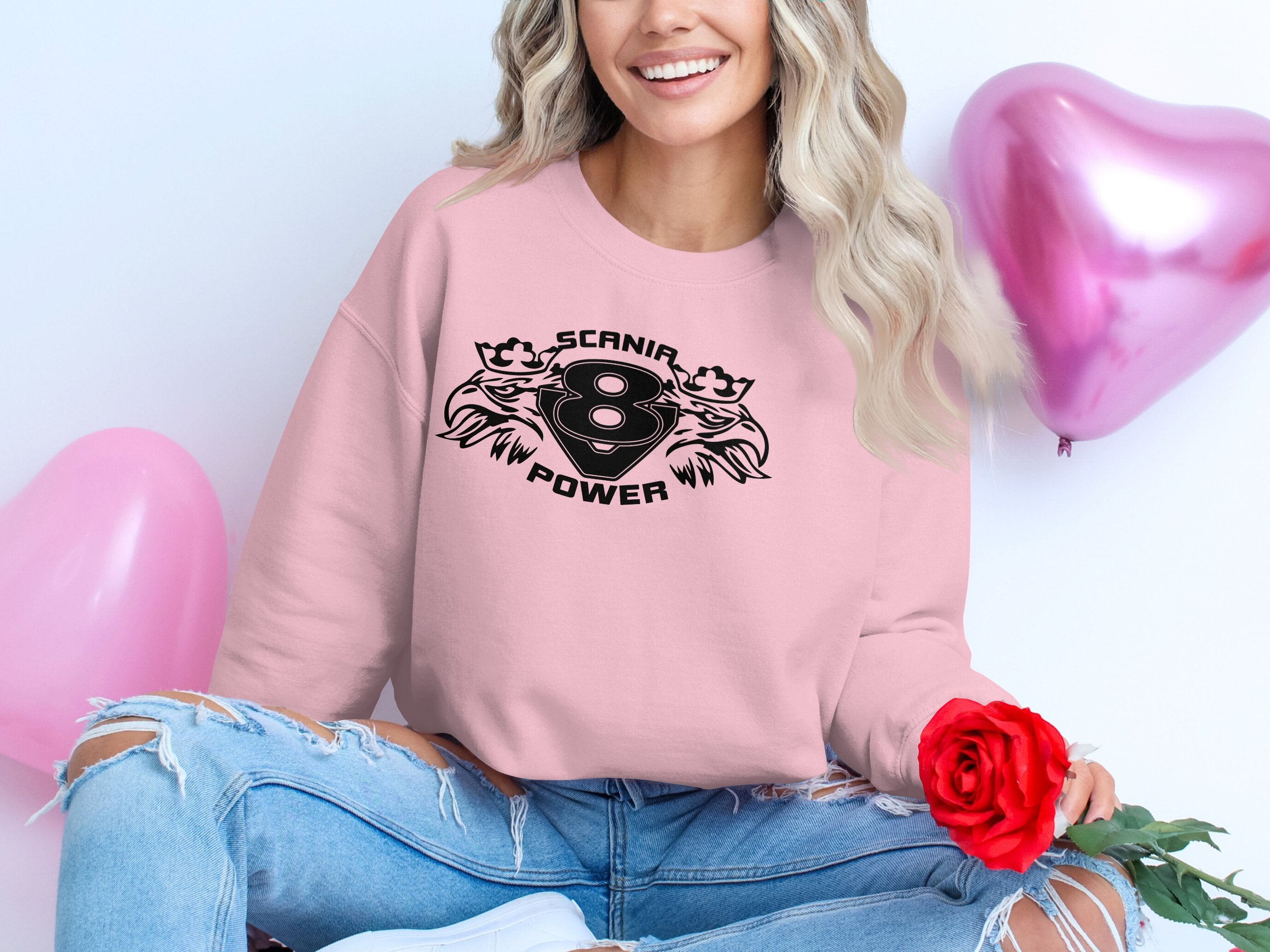 A person sits stylishly cross-legged on the floor, wearing a pink Garment Graphics Scania Power V8 sweatshirt with distressed jeans while holding a red rose. Pink heart-shaped balloons float in the background, complementing their cool printed attire.