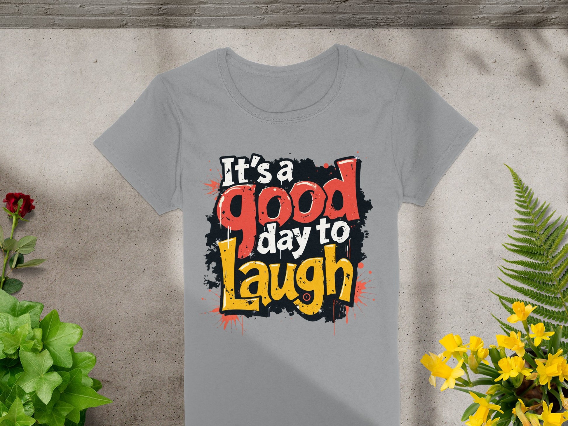 Garment Graphics presents a semi-fitted gray T-shirt featuring Its a good day to Laugh in vibrant white, red, and yellow letters on a black splash background. The cheerful design is displayed on concrete with lush green and yellow plants around it.