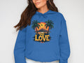 A person in a distinctive hoodie from Garment Graphics, adorned with a tropical sunset scene of palm trees and mountains, smiles slightly. ONE LOVE is boldly emblazoned below the image, highlighting their unique style as their long dark hair frames their face.