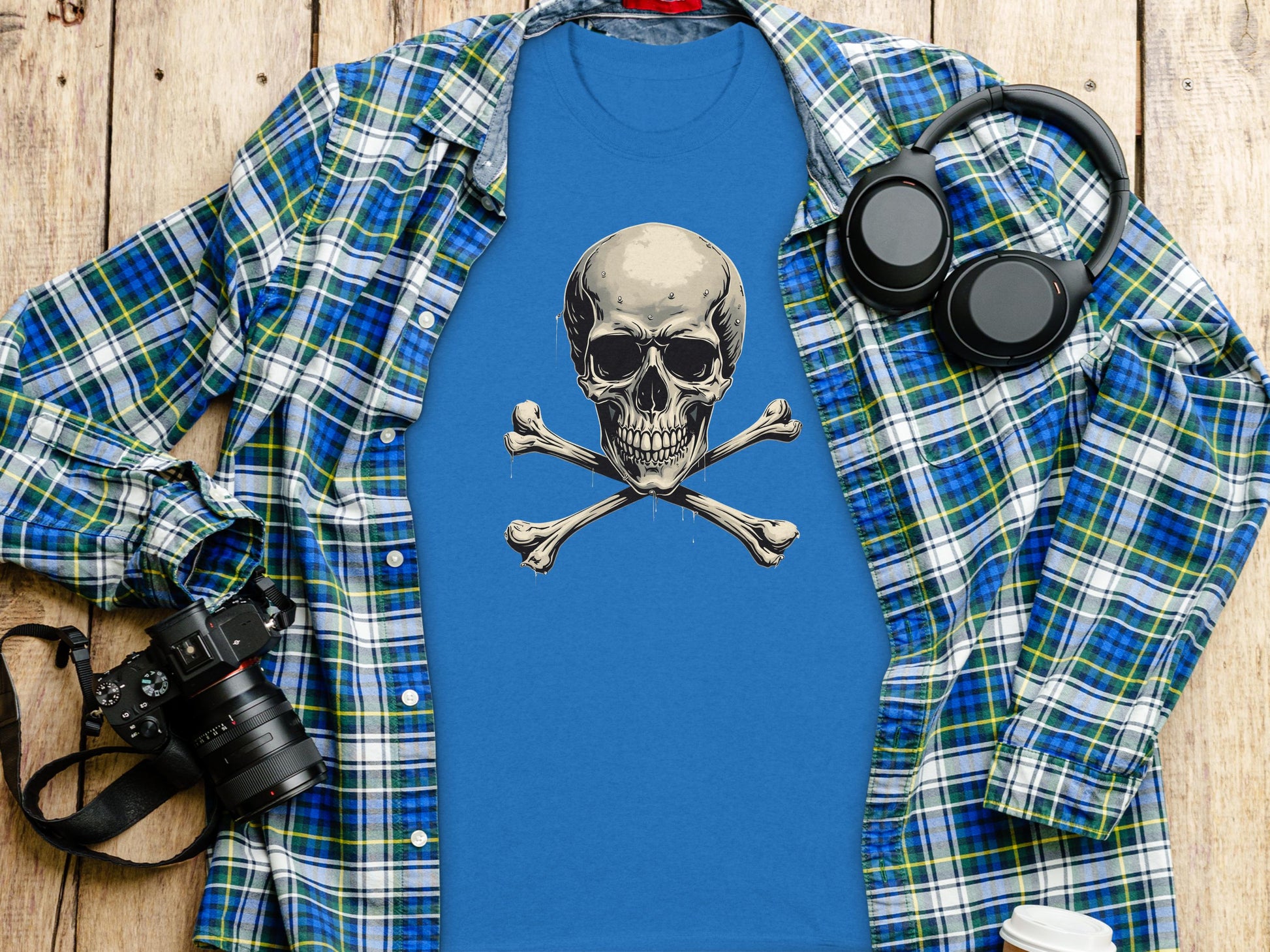A Garment Graphics graphic tee with a bold skull and crossbones design lies flat on a wooden surface, making a statement. Its paired with a blue and green plaid shirt, surrounded by a camera, headphones, and coffee cup.