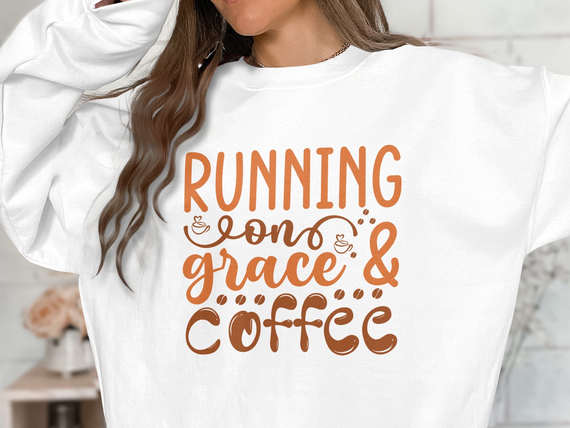 Someone is wearing a Garment Graphics unisex graphic sweatshirt with Running on Grace and Coffee in brown letters. The design features charming coffee cup illustrations, and their hair partially hides their face.