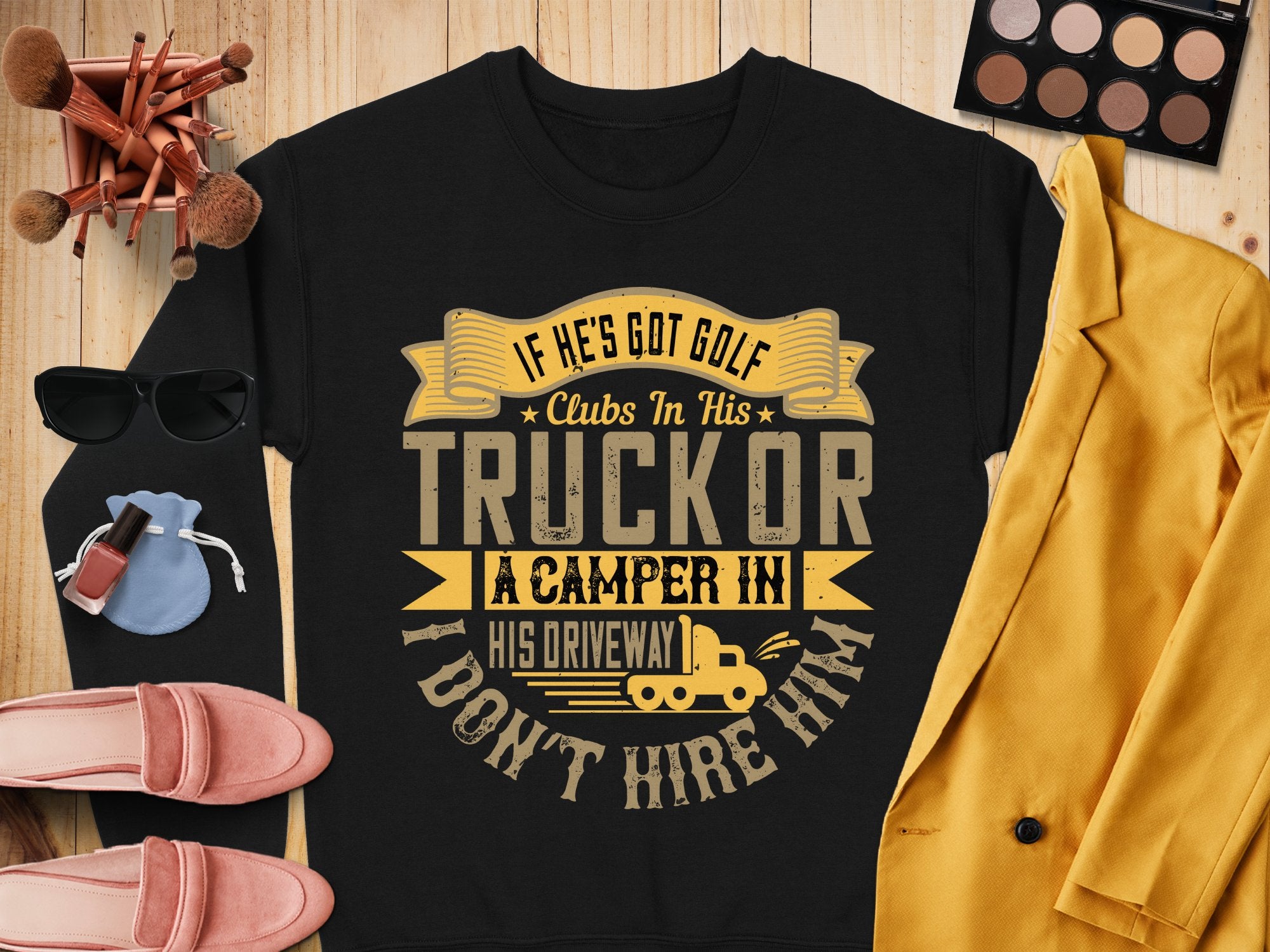 A black Garment Graphics sweatshirt humorously displays: If hes got golf clubs in his truck or a camper in his driveway, I dont hire him. Styled with sunglasses, makeup, a mustard blazer, pink shoes, and a blue pouch on wood for a perfect casual look.