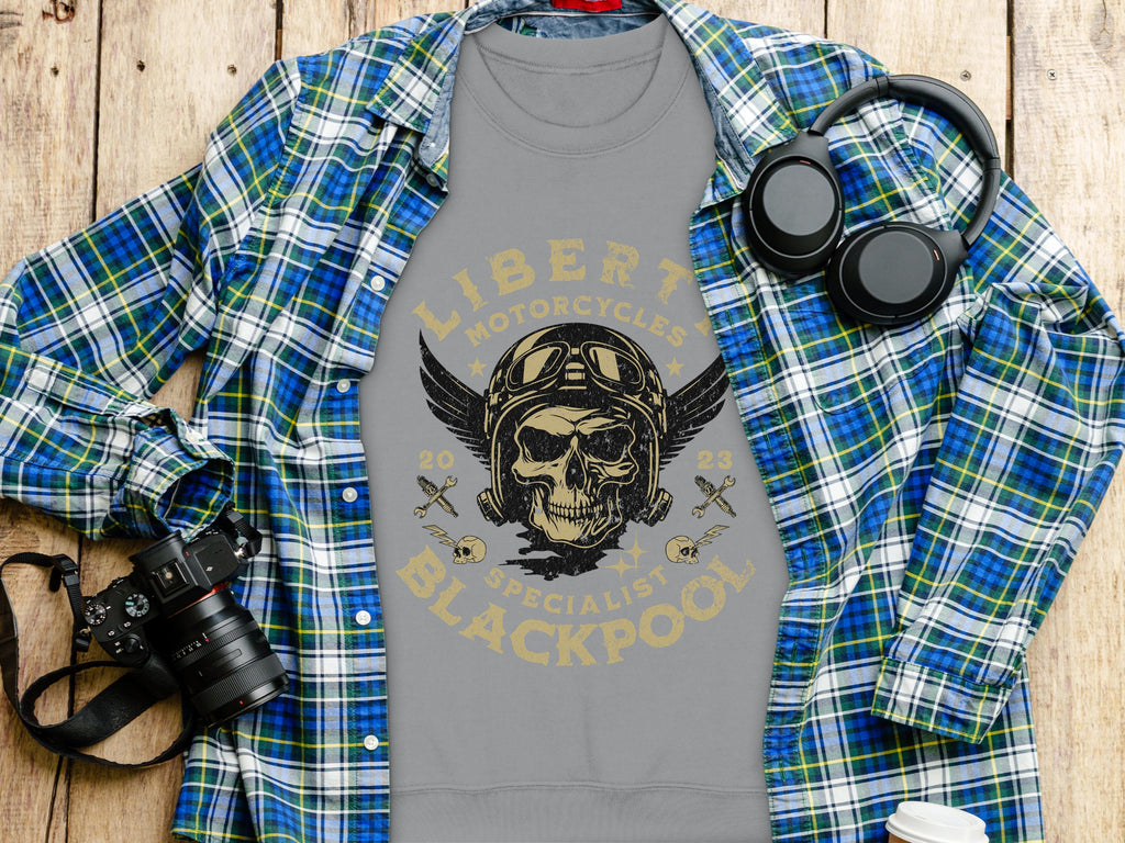 A gray T-shirt from Garment Graphics featuring a skull, wings, goggles, and motorcycle theme with Liberty Motorcycles Specialist Blackpool 2023 is laid on a wooden surface. Next to it are a blue plaid shirt, headphones, and a camera.