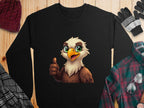 A black cartoon eagle sweatshirt from Garment Graphics with a cheerful design is displayed on wood. Its surrounded by a maroon knit hat, black and gray gloves, a black and white plaid scarf, and a green and red flannel shirt. The unisex sizing ensures versatility for any wardrobe.