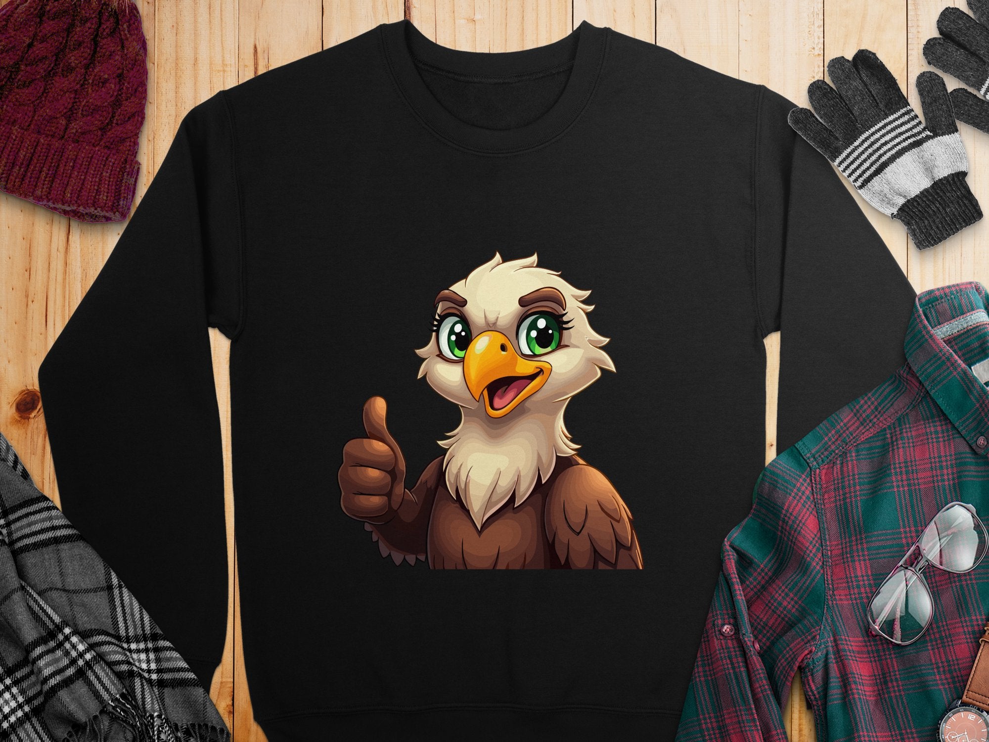 A black cartoon eagle sweatshirt from Garment Graphics with a cheerful design is displayed on wood. Its surrounded by a maroon knit hat, black and gray gloves, a black and white plaid scarf, and a green and red flannel shirt. The unisex sizing ensures versatility for any wardrobe.