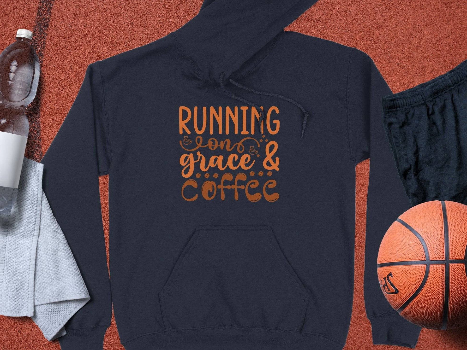 A Garment Graphics dark, medium-heavy fabric hoodie with Running on grace & coffee in orange sits on a sports court alongside a basketball, black shorts, a white towel, and a clear water bottle.