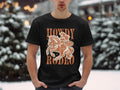 Amidst snow-covered trees, an enthusiast stands outdoors in a black Garment Graphics Rodeo T-shirt, featuring a cowboy on horseback with the bold statement Howdy Rodeo and a smaller circle saying Not Sorry.