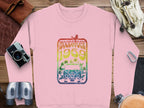 A Garment Graphics pink sweatshirt adds retro flair with Woodstock 1969 above a flower-adorned vintage van, surrounded by a camera, binoculars, book, bag, and leather accessories on wood.