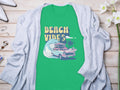 The Garment Graphics green graphic T-shirt showcases Beach Vibes with a classic car, surfboards, waves, and palm trees. Its laid on light wood and partially covered by a gray cardigan, surrounded by shoes and flowers.