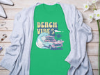 The Garment Graphics green graphic T-shirt showcases Beach Vibes with a classic car, surfboards, waves, and palm trees. Its laid on light wood and partially covered by a gray cardigan, surrounded by shoes and flowers.