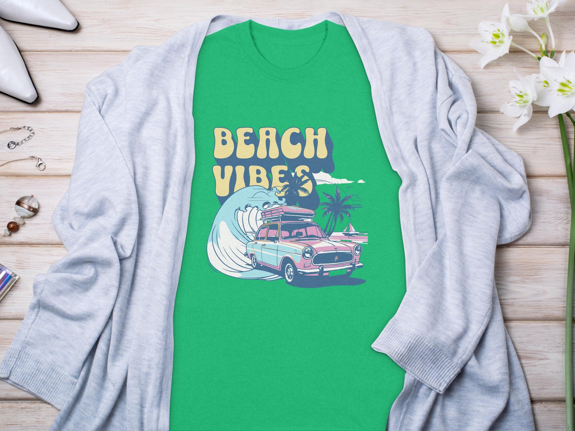 The Garment Graphics green graphic T-shirt showcases Beach Vibes with a classic car, surfboards, waves, and palm trees. Its laid on light wood and partially covered by a gray cardigan, surrounded by shoes and flowers.
