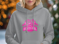 A person wearing a Garment Graphics Always Party Beach Girl hoodie flaunts a vibrant design with silhouettes of three people and palm trees. The casual style stands out in gray with a pink front print, set against a blurred outdoor backdrop.