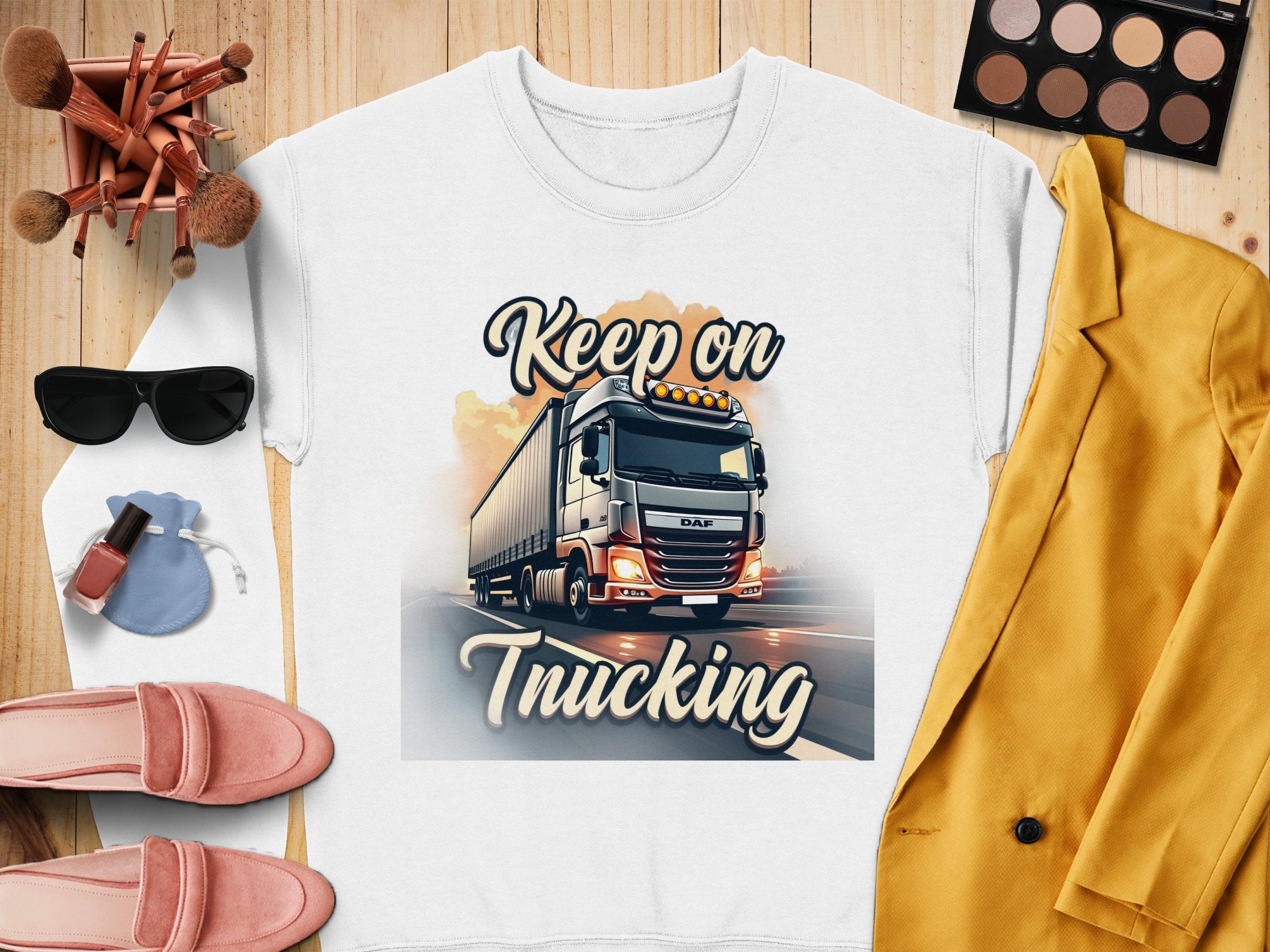 Garment Graphics unisex graphic sweatshirt, Keep on Trucking, is styled in a flat lay with makeup brushes, a palette, sunglasses, lipstick, pink loafers, a small pouch, and a yellow blazer on a wooden surface.