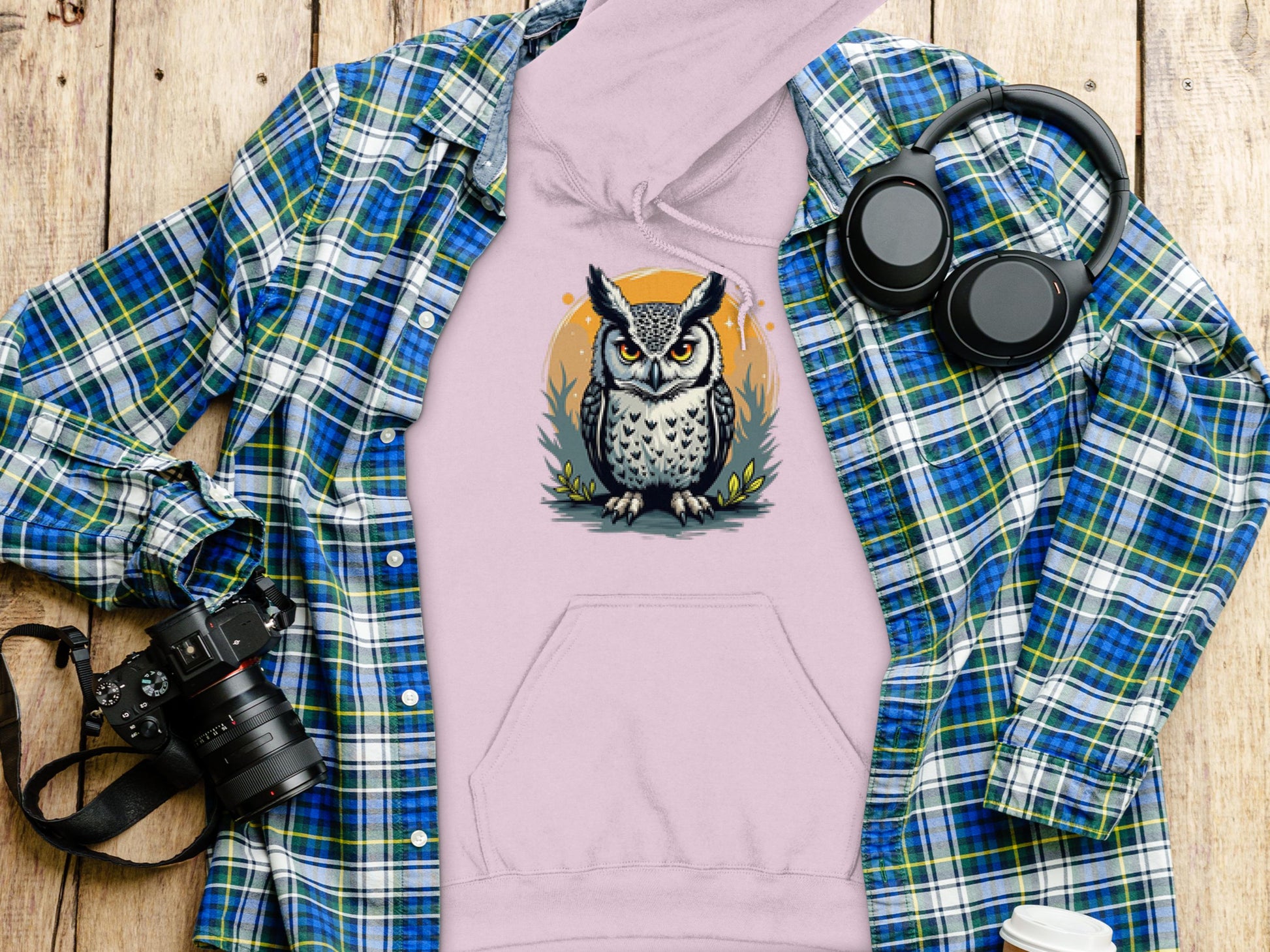 A Garment Graphics light pink hoodie with a fierce owl design is on a wooden surface, accompanied by a blue-green plaid shirt, black headphones, a camera, and a white lidded cup—each enhancing the distinct style ensemble.