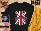 A flat lay showcases a black unisex sweatshirt from Garment Graphics featuring the British flag and the text Dont Blame Me, I Didnt Vote For Him, surrounded by sunglasses, pink loafers, a makeup bag, brushes, a makeup palette, nail polish, and a yellow jacket.