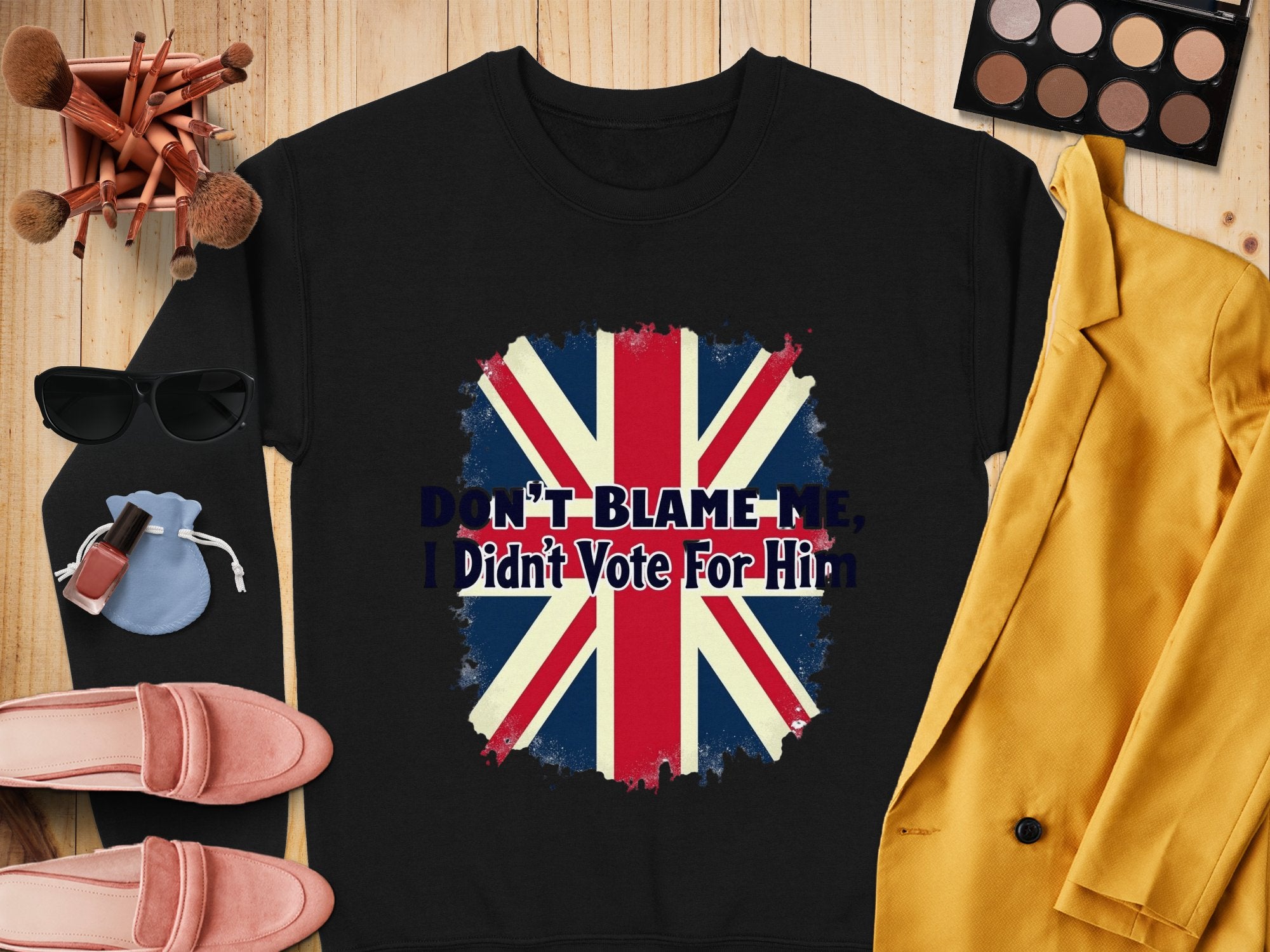 A flat lay showcases a black unisex sweatshirt from Garment Graphics featuring the British flag and the text Dont Blame Me, I Didnt Vote For Him, surrounded by sunglasses, pink loafers, a makeup bag, brushes, a makeup palette, nail polish, and a yellow jacket.