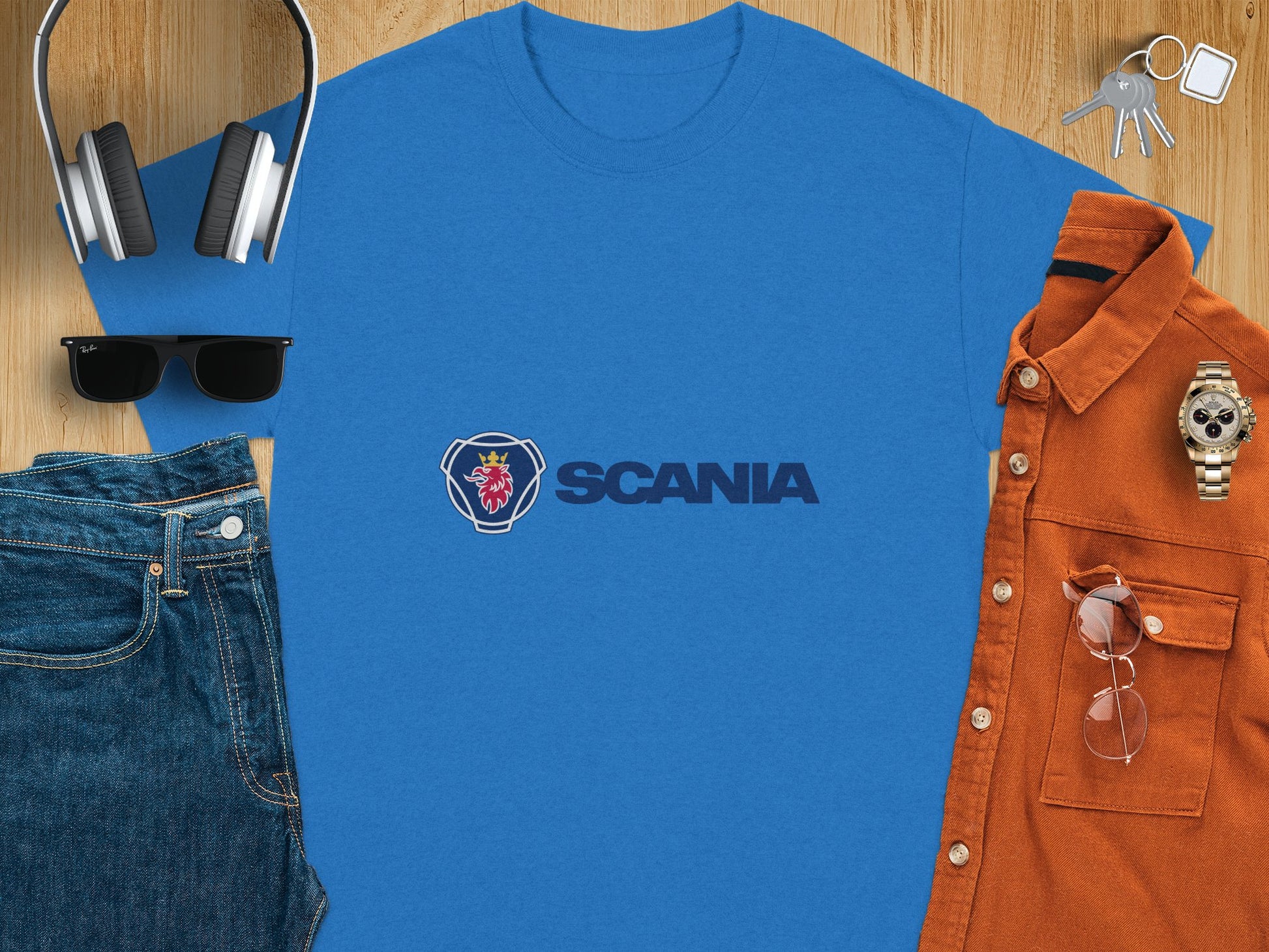 On a wooden surface, theres a blue Garment Graphics Scania Logo T-shirt, headphones, sunglasses, jeans, a brown button-up shirt, a watch, keys, and a smartphone. This neatly arranged flat lay highlights stylish Scania merchandise.
