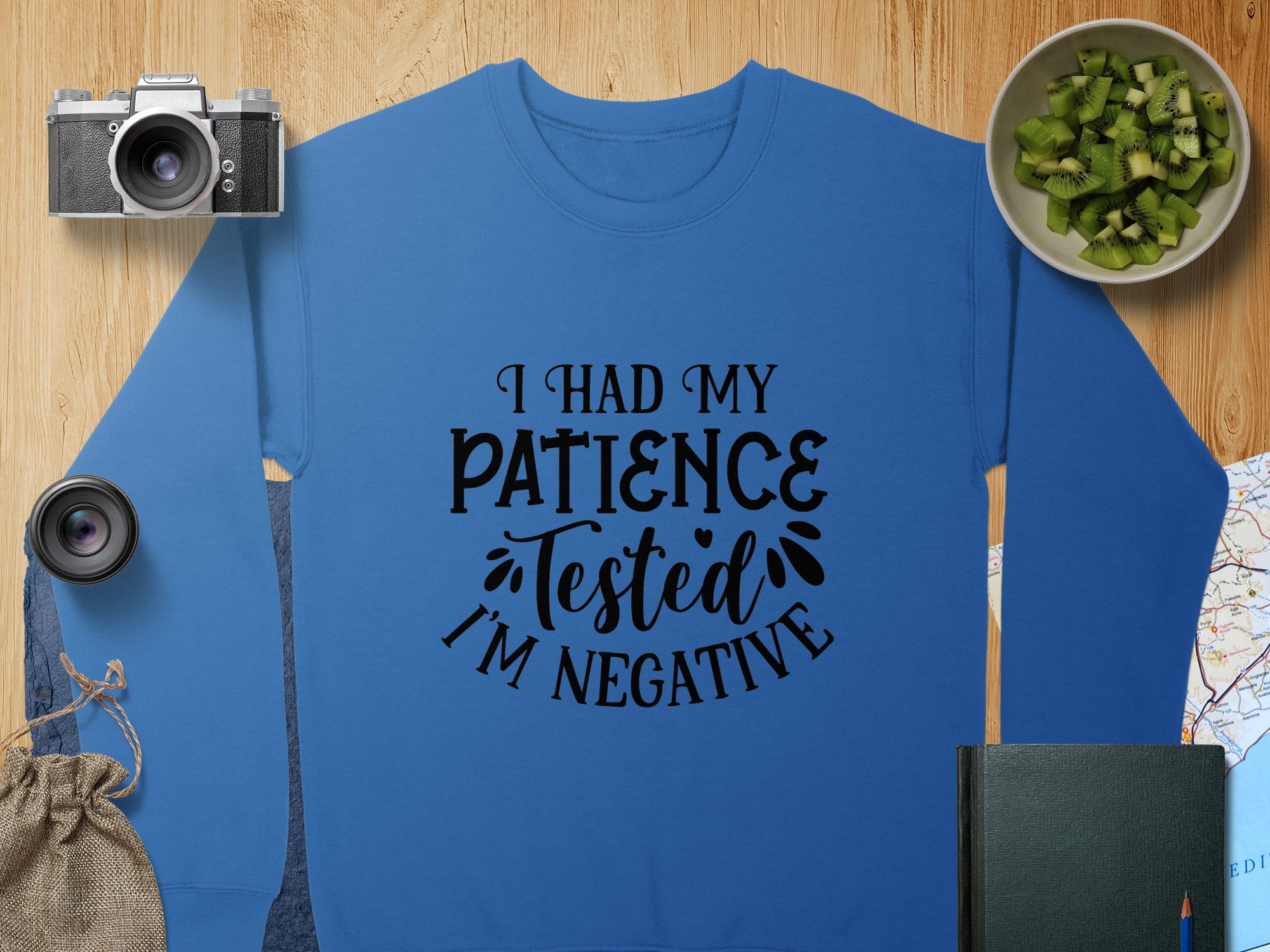 The quirky blue Garment Graphics sweatshirt features I had my patience tested, Im negative in black, ideal for casual outings. Its surrounded by a camera, lens, fruit bowl, jute pouch, book, and a partly visible map on wood. Unisex sizing offers versatility.