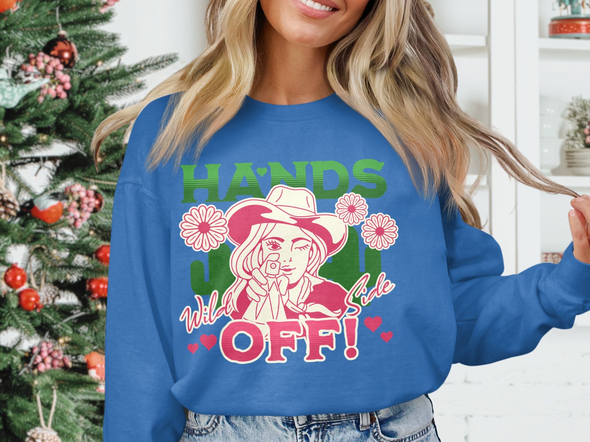 A person wearing a blue Garment Graphics Hands Off Wild Side Sweatshirt stands by a Christmas tree, featuring a cowboy graphic with flowers and hearts. Its loose fit makes it perfect for the holidays.