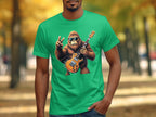 A person in a Garment Graphics green T-shirt with a cartoon Bigfoot playing an electric guitar and making a peace sign stands on an autumn-leaf-lined path, an ideal scene for music lovers.