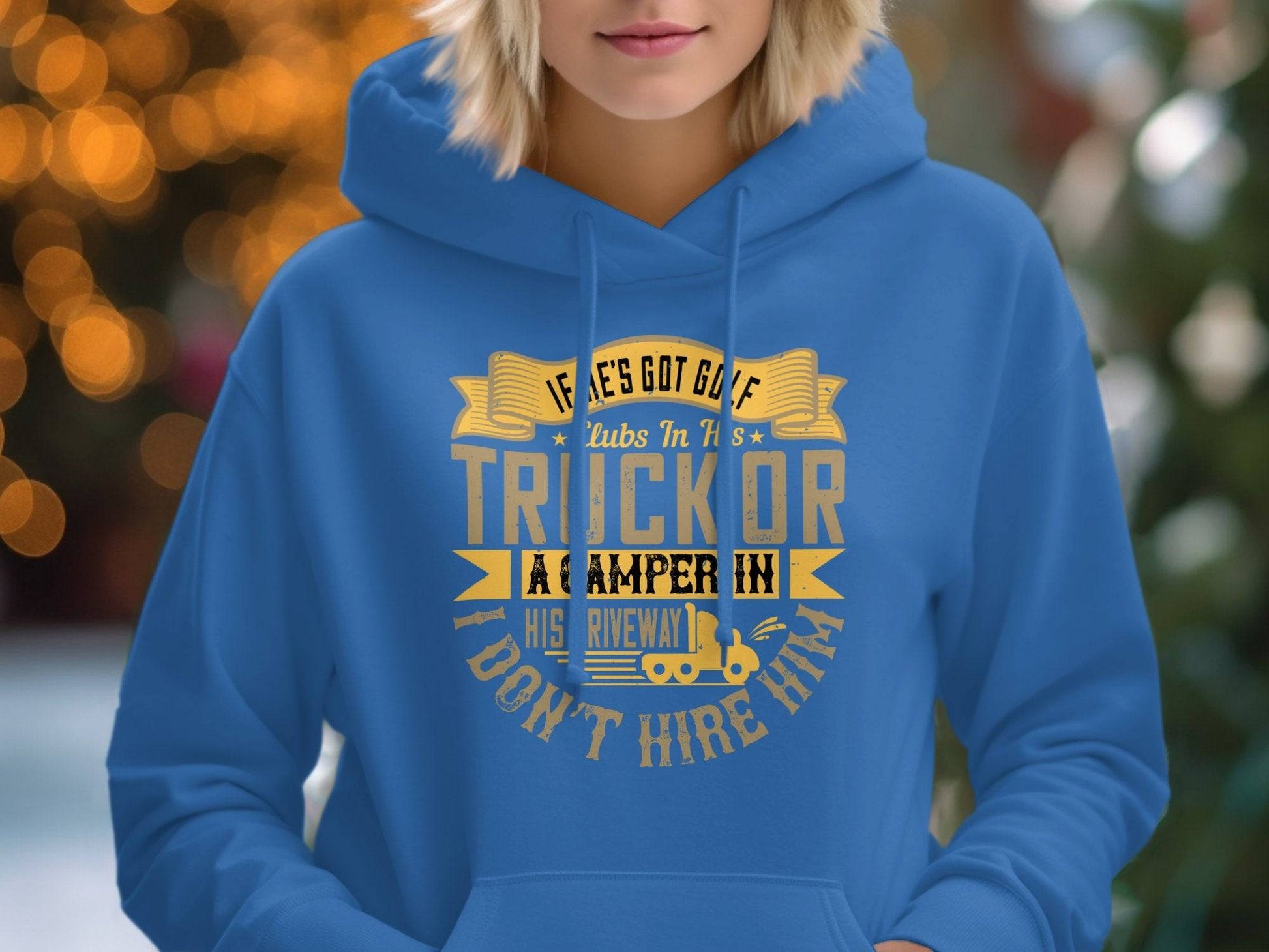 A person dons a blue golf hoodie from Garment Graphics with the witty phrase, If hes got golf clubs in his truck or a camper in his driveway, I dont hire him. The softly blurred background sparkles with glowing lights.