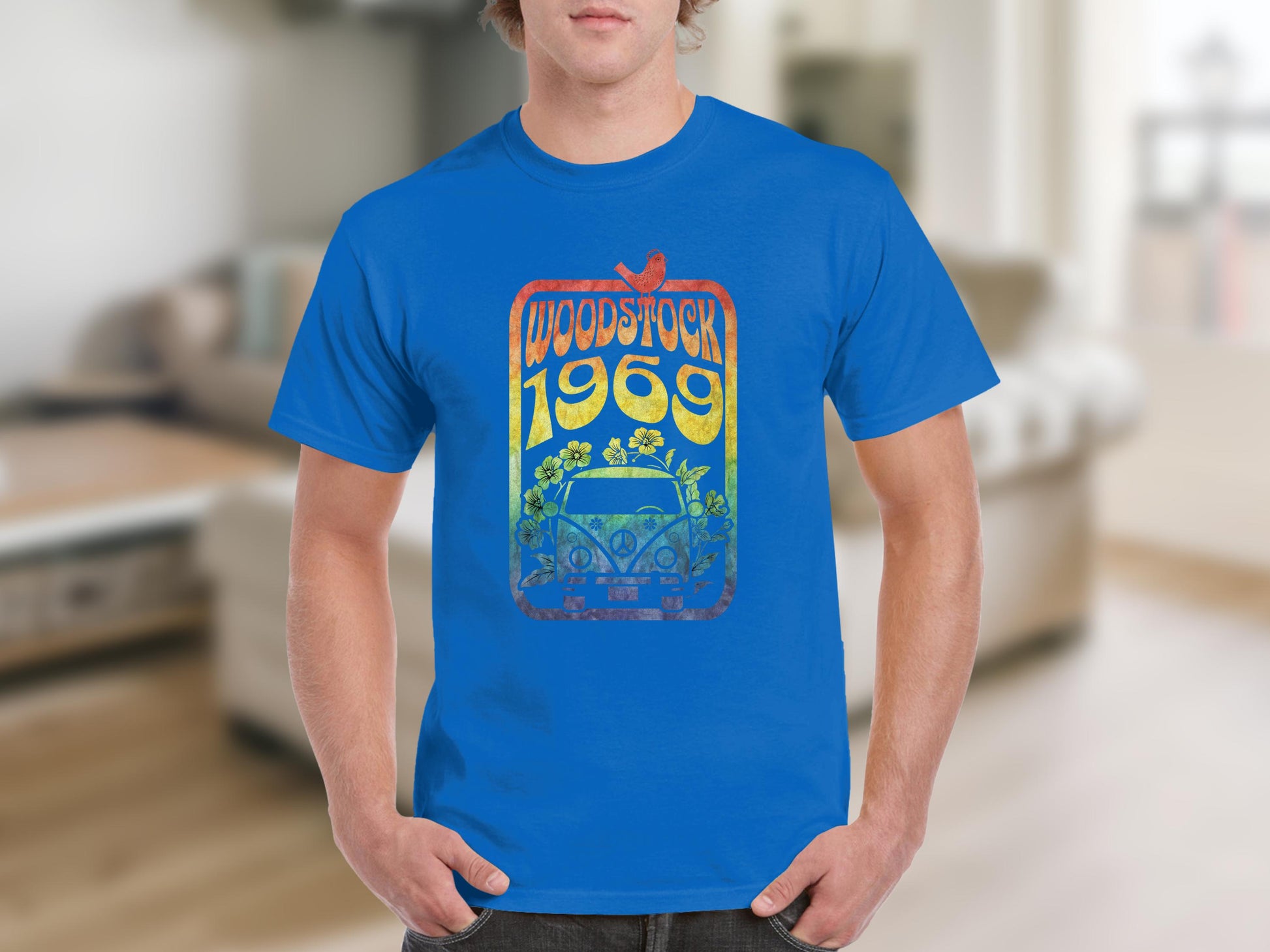 A person wearing a Garment Graphics blue T-shirt displays a vibrant Woodstock 1969 design with a retro van and flowers, ideal for music lovers, against a softly blurred interior background.