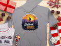 A cozy Garment Graphics gray hiking hoodie showcases a hiker silhouette and the text Retired 2025 - Not My Problem Anymore, featuring a sunset mountain design, surrounded by wrapped gifts, a sweater, candy canes, and festive decorations on a wooden surface.