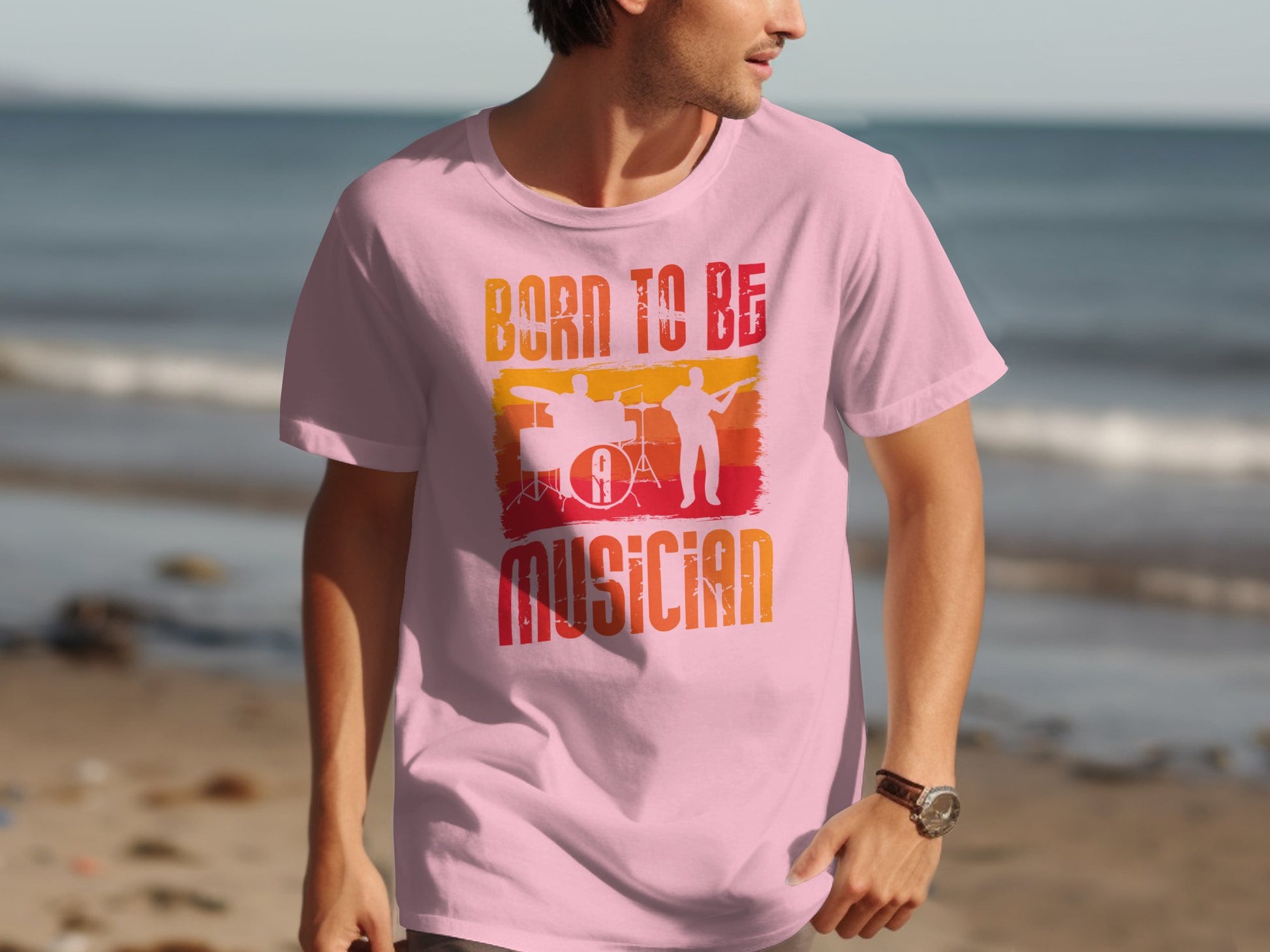 A man walks on the beach wearing a Garment Graphics light pink Born To Be Musician T-shirt, showcasing drummer and guitarist silhouettes. The shirt boldly states BORN TO BE A MUSICIAN in vibrant letters, with the ocean calmingly in the background—a music lovers ideal scene.