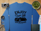 A Garment Graphics blue sweatshirt, featuring a vintage ride graphic and the words Enjoy Classic Car, Vintage Ride Community, Aircooled, is laid out on a wooden surface surrounded by essentials: a camera, lens, map, notebook, and a bowl of kiwi slices.