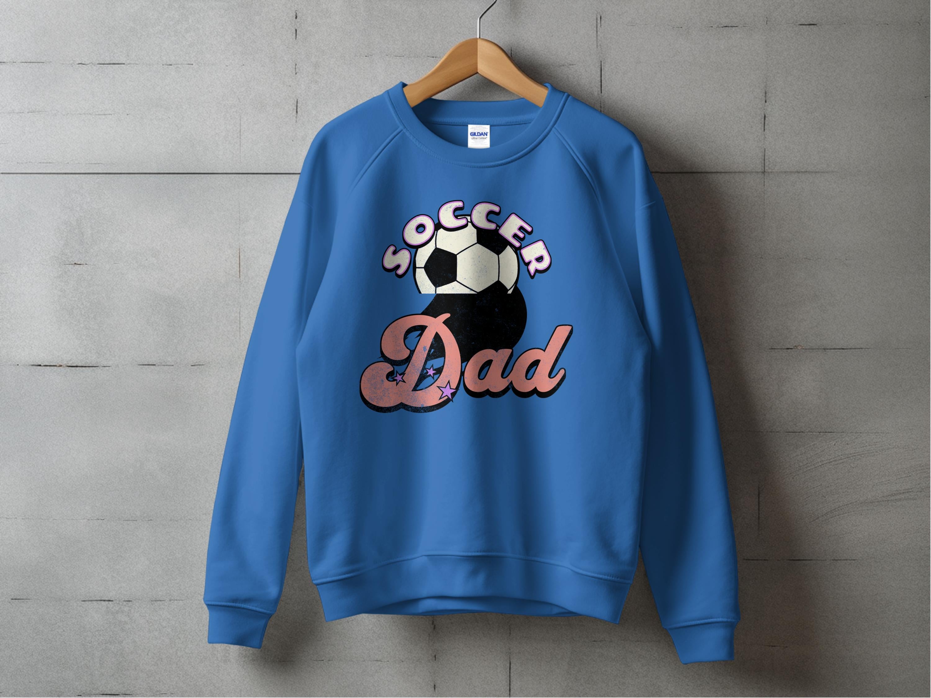 A Garment Graphics sweatshirt with a Soccer Dad design, featuring a soccer ball graphic between the words, hangs on a wooden hanger against a gray concrete wall.