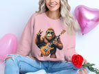 Sporting long wavy hair, a person dons a Garment Graphics pink sweatshirt depicting a guitar-playing gorilla. They hold a red rose, sitting beside pink heart-shaped balloons and smiling while wearing ripped jeans.