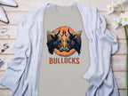 The Classic Fit light gray t-shirt from Garment Graphics Bullocks Pintable T-Shirts collection features two bulls facing each other against an orange circle backdrop with BULLOCKS below. Displayed on a wooden surface beside a light gray cardigan, its perfect for Family Sizing adventures.