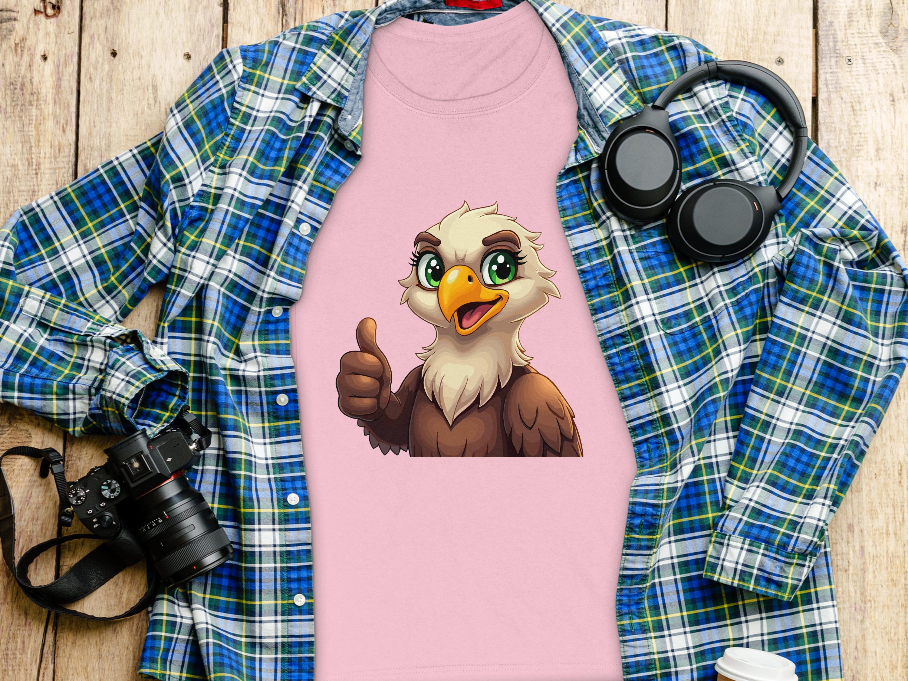 The Garment Graphics pink cartoon T-shirt with an eagle giving a thumbs up, placed on a plaid shirt, is perfect for animal lovers and is surrounded by a camera, headphones, and cup on a wooden surface.