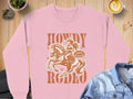 Garment Graphics offers a pink graphic print sweatshirt ideal for casual wear, showcasing a cowgirl on horseback with Howdy Rodeo and Not a Beginner. Styled with a denim jacket and latte art on a wooden table.