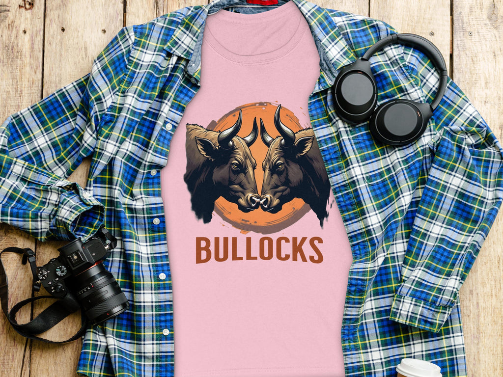 A Garment Graphics pink T-shirt with bold bull illustrations and BULLOCKS is laid over a plaid shirt. Nearby, a camera, black headphones, and a white bottle are artfully arranged on a wooden surface.