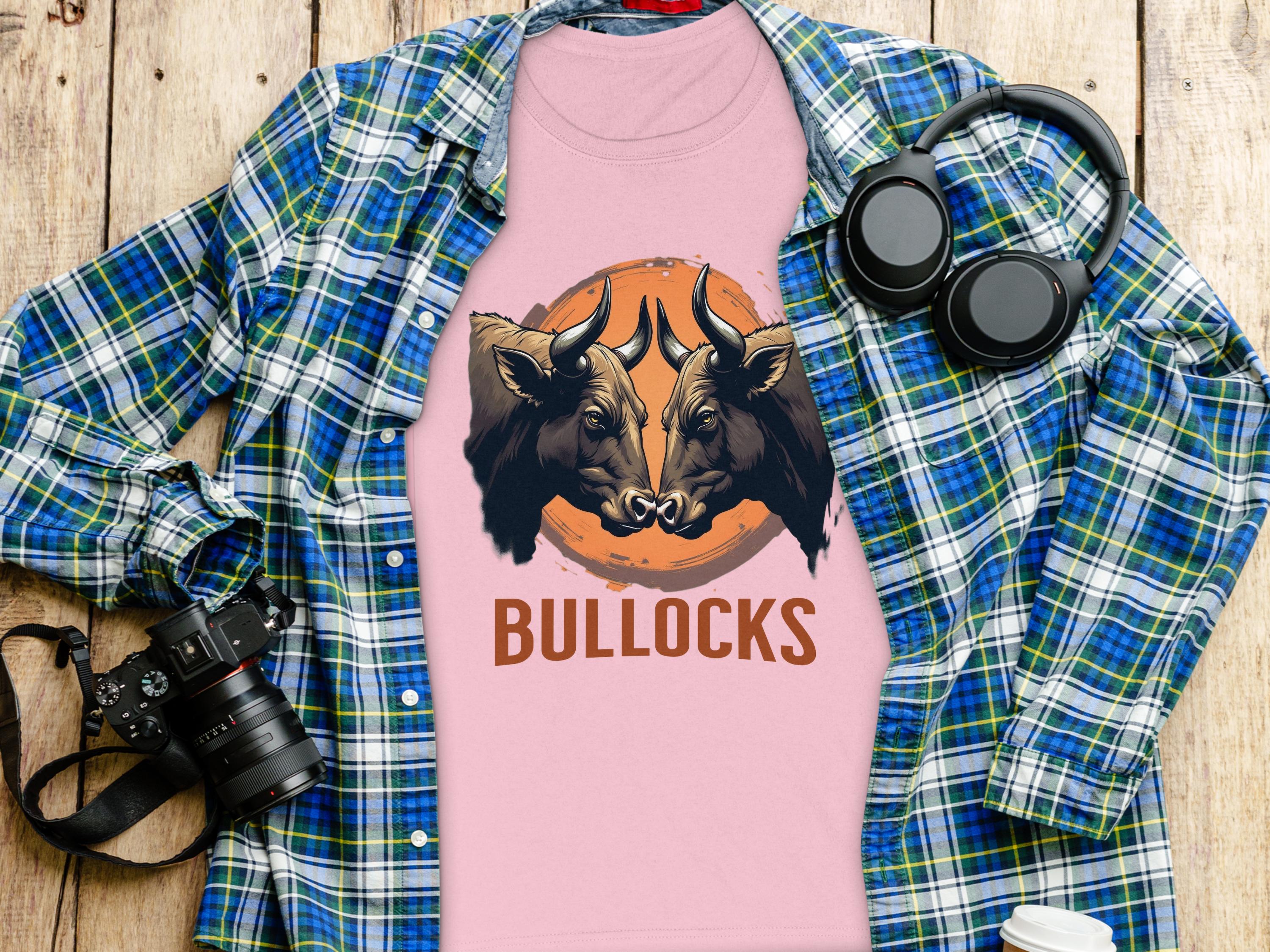 A Garment Graphics pink T-shirt with bold bull illustrations and BULLOCKS is laid over a plaid shirt. Nearby, a camera, black headphones, and a white bottle are artfully arranged on a wooden surface.
