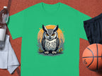 A Garment Graphics green T-shirt features an owl graphic in front of a yellow circle, exuding artistic flair. It rests on a reddish surface and is surrounded by a clear plastic water bottle, black shorts, a basketball, and a gray towel.